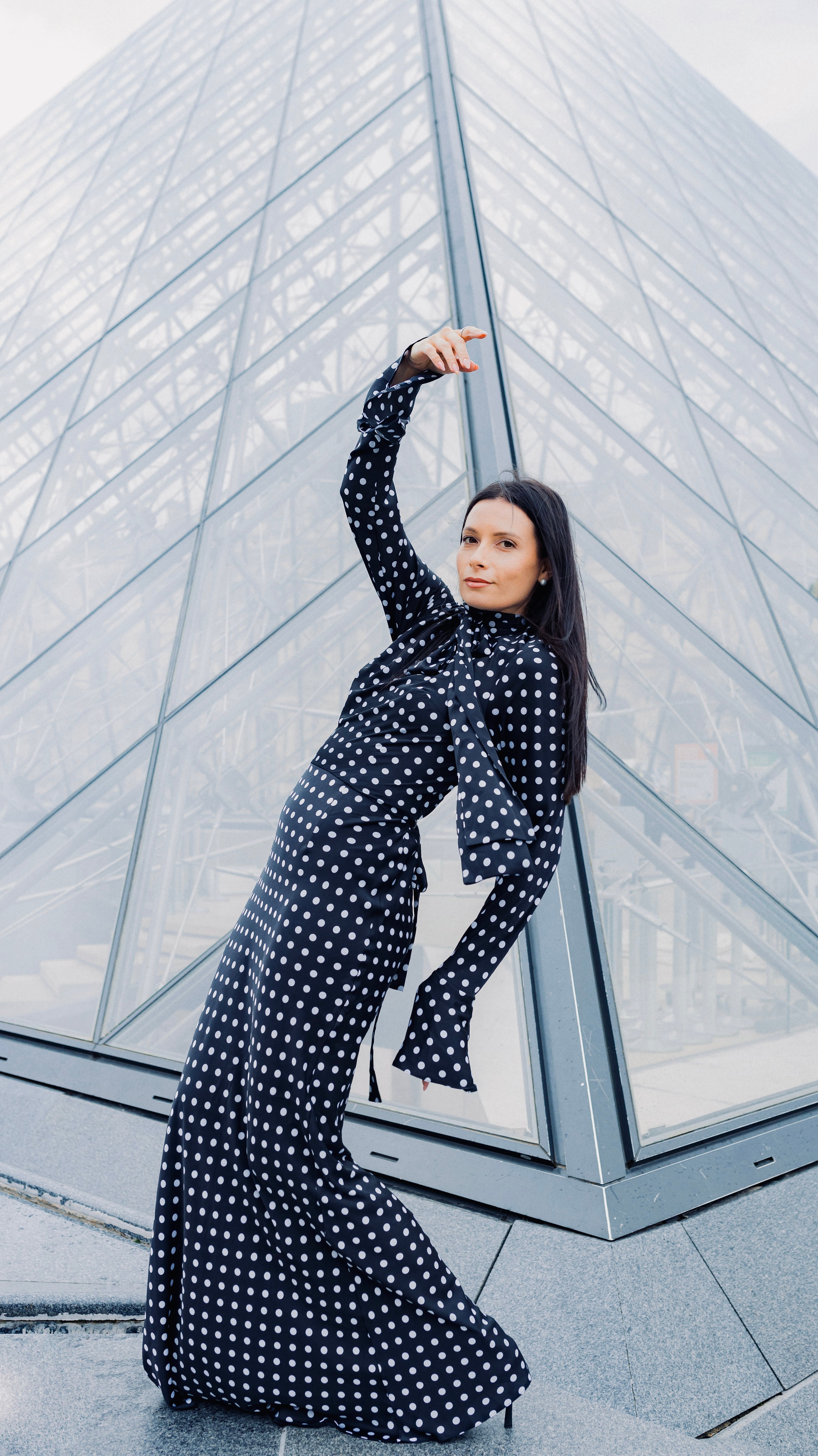 Woman in a long, polka-dot dress striking a sculptural pose in front of the Louvre pyramid